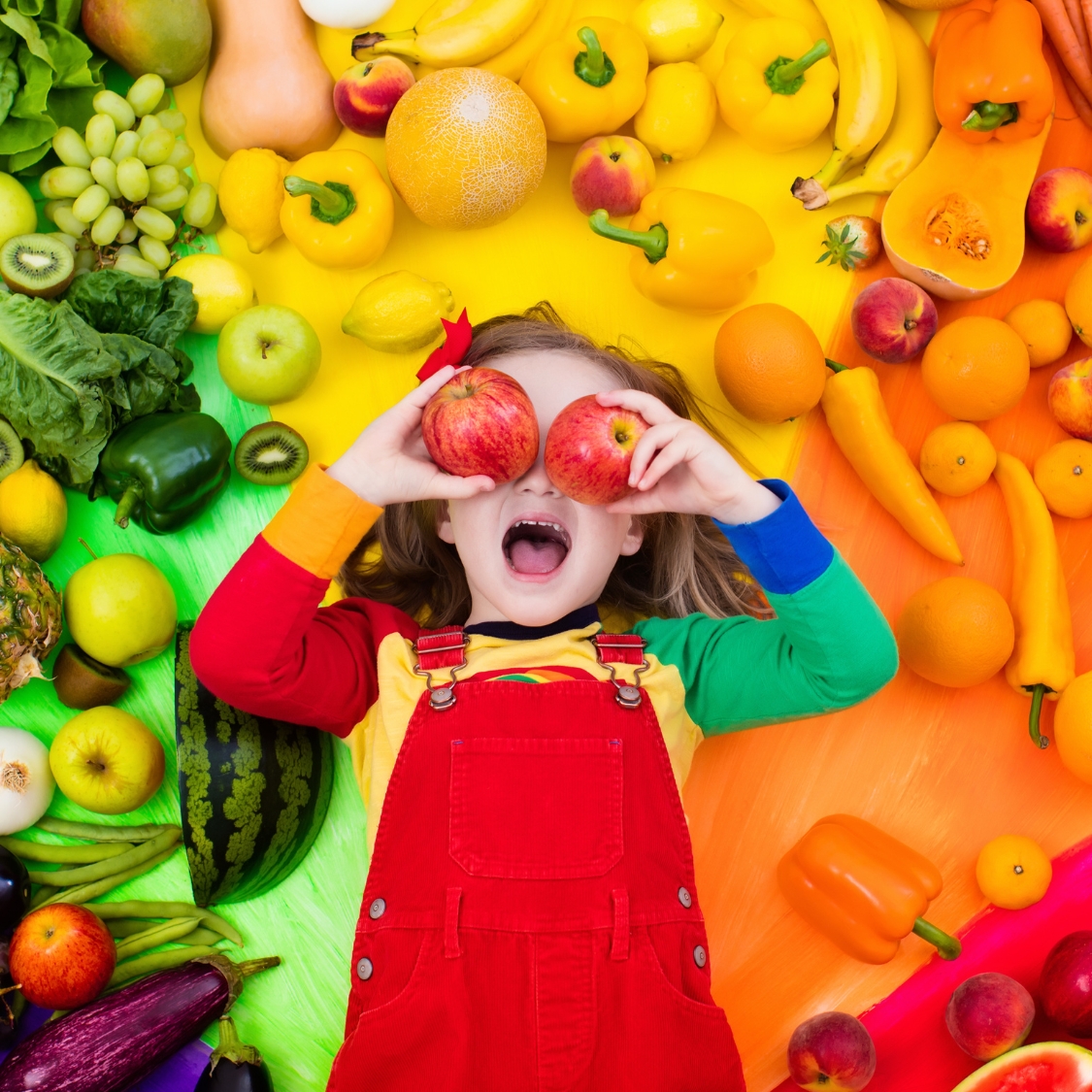 A young smiling girl, surrounded by healthy fruits and vegetables, holds two apples in front of her eyes.