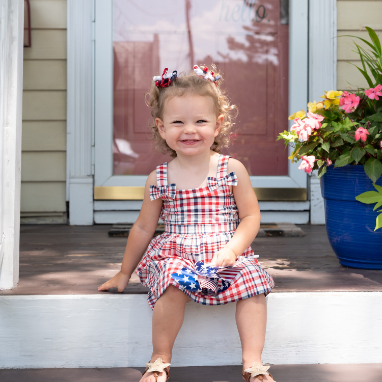 May-blog-2 Young child in a red, white and blue dress. She's sitting on the front steps and holds an american flag pinwheel.