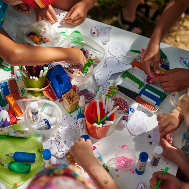 A group of diverse kids gathered around a table outside. The table is covered in fun craft items